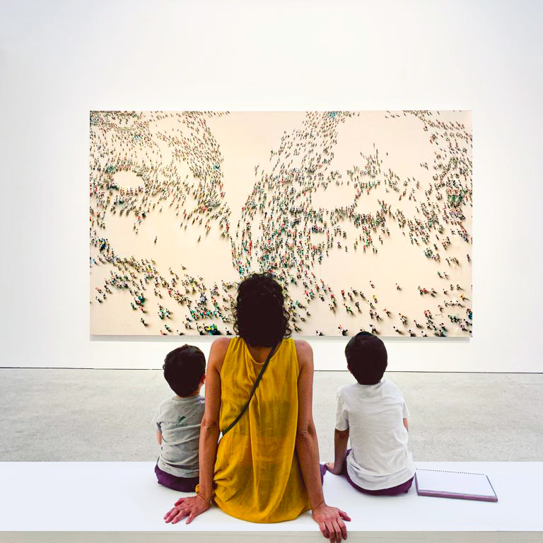 Vista posterior de una mujer adulta y dos niños sentados en un banco, observando un cuadro en un museo.